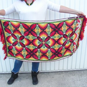 May include: A rectangular, colorful textile with a floral pattern. The design features red, orange, yellow, and green hues. The edges are trimmed with a green border and orange fringe.