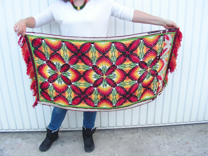 May include: A rectangular, colorful textile with a floral pattern. The design features red, orange, yellow, and green hues. The edges are trimmed with a green border and orange fringe.