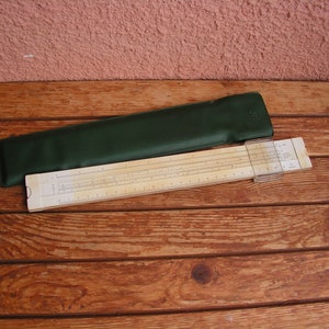 Może przedstawiać: Wzorcowa suwmiarka z zielonym skórzanym etui. Suwmiarka wykonana jest z drewna i ma białe tło z czarnymi oznaczeniami. Etui posiada metalowe zapięcie.