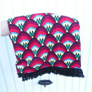 May include: A vibrant, hand-stitched textile with a repeating fan-shaped pattern in shades of red, yellow, blue, and green. The edges are finished with a black fringe. The textile is held against a white background.