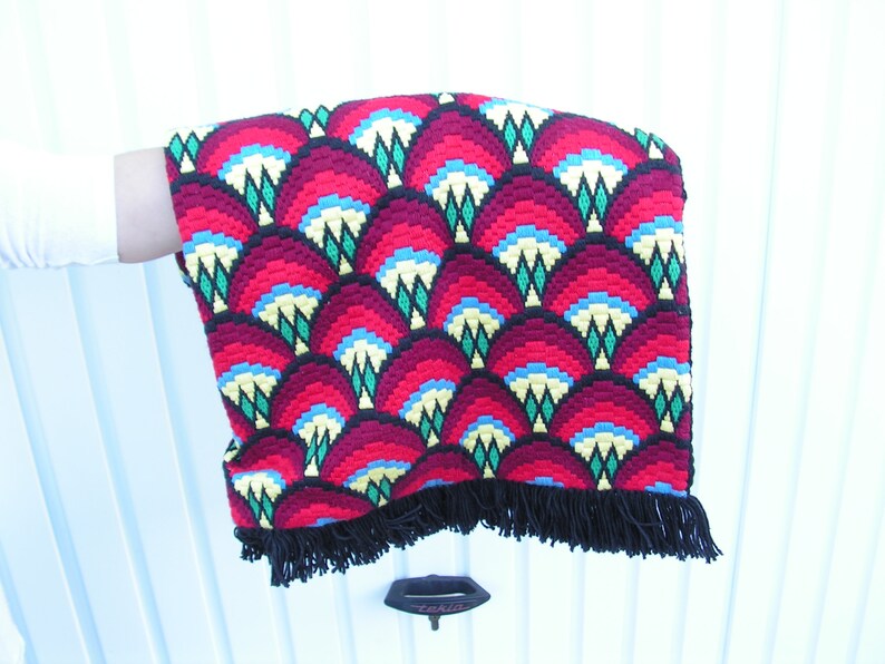 May include: A vibrant, hand-stitched textile with a repeating fan-shaped pattern in shades of red, yellow, blue, and green. The edges are finished with a black fringe. The textile is held against a white background.