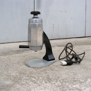 May include: A silver and black electric stovetop espresso maker with a cord and plug. The maker has a round base and a tall, octagonal-shaped pot.