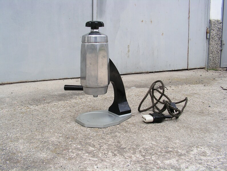 May include: A silver and black electric stovetop espresso maker with a cord and plug. The maker has a round base and a tall, octagonal-shaped pot.