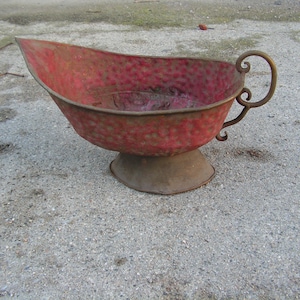 May include: A red metal planter with a decorative handle and a raised base. The planter is made of a textured metal and has a worn, rustic look.
