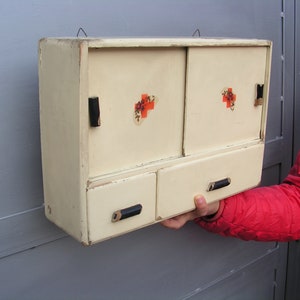 May include: A vintage white wooden medicine cabinet with two doors and a drawer. The doors have red crosses with floral accents. The cabinet has black metal handles.