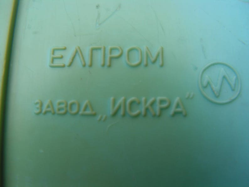 May include: A close-up of a green plastic object with the text "EЛПРOМ ЗАВОД, ИСКРА" and a symbol of a lightning bolt in a circle.