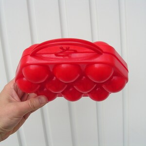 May include: A red plastic egg carton with a handle. The carton has 12 individual compartments for holding eggs.