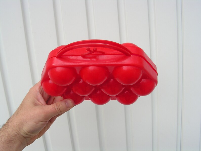 May include: A red plastic egg carton with a handle. The carton has 12 individual compartments for holding eggs.