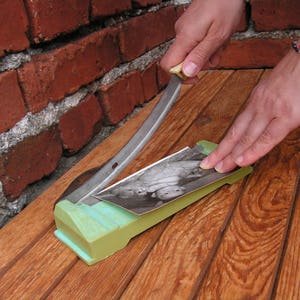 May include: A person is using a vintage paper cutter to cut a black and white photograph of two children. The cutter has a green handle and a silver blade. The photograph is on a wooden surface.