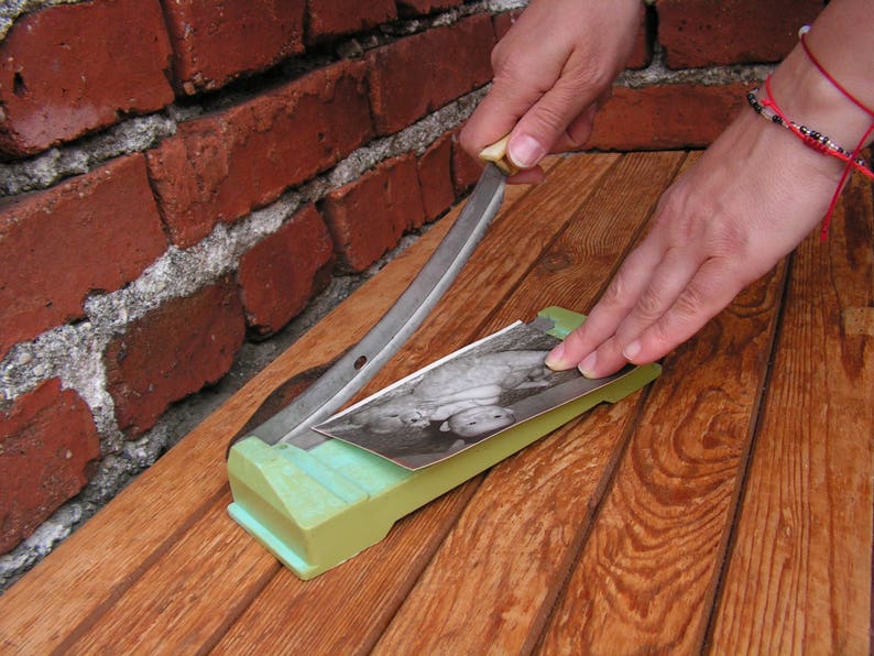 May include: A person is using a vintage paper cutter to cut a black and white photograph of two children. The cutter has a green handle and a silver blade. The photograph is on a wooden surface.