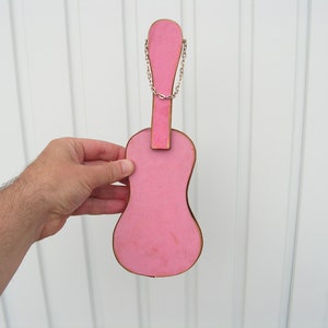 May include: A pink wooden guitar shaped ornament with a silver chain attached to the top. The ornament is hanging on a white wall.
