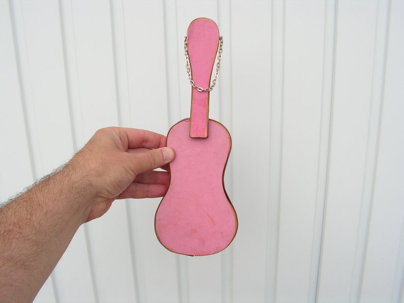 May include: A pink wooden guitar shaped ornament with a silver chain attached to the top. The ornament is hanging on a white wall.