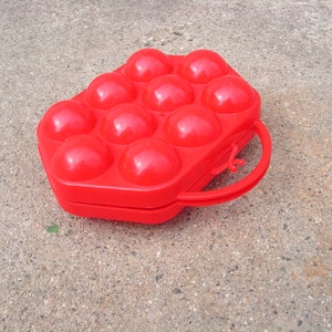 May include: A red plastic egg carton with a handle. The carton has 12 individual compartments for holding eggs.