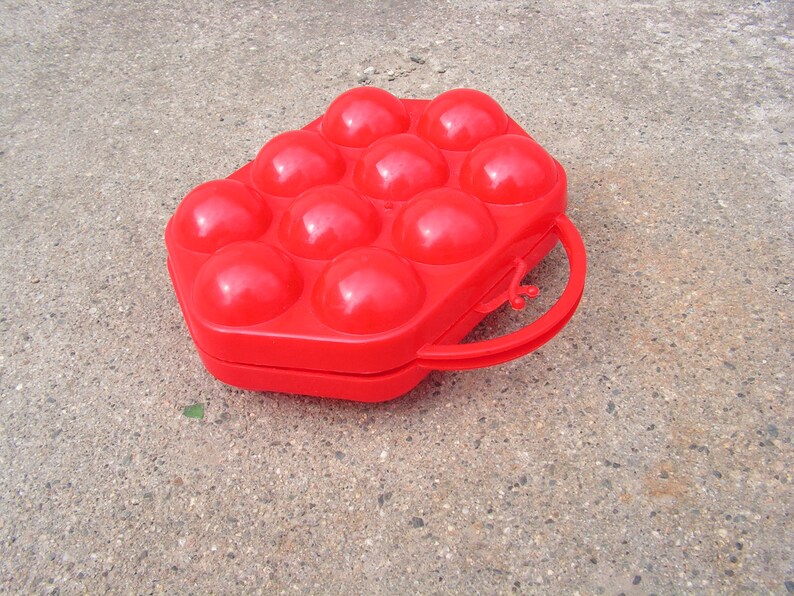 May include: A red plastic egg carton with a handle. The carton has 12 individual compartments for holding eggs.