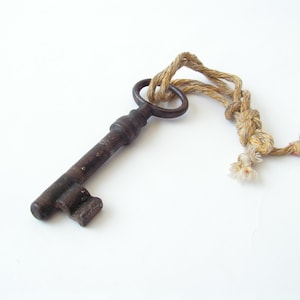 May include: An antique, dark brown metal key with a circular handle and a decorative bit. The key is attached to a twisted rope with a knot and frayed ends. The background is plain white.
