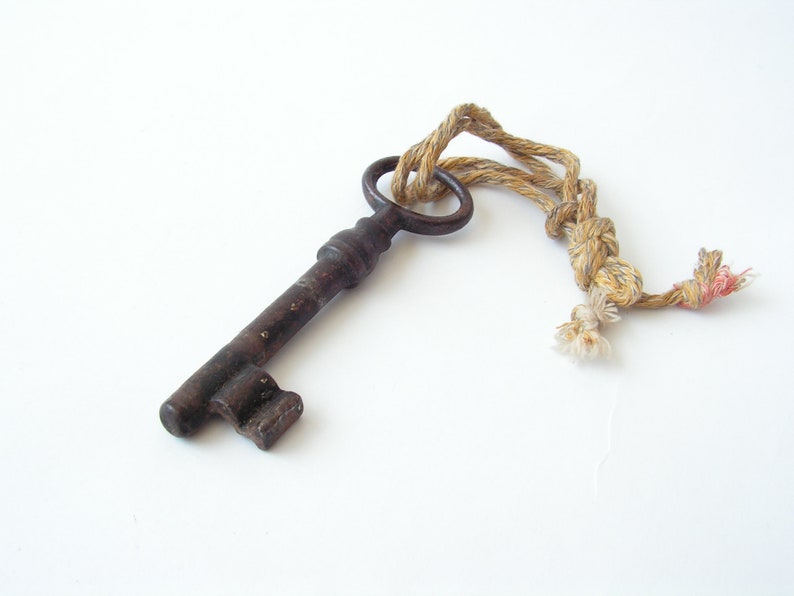 May include: An antique, dark brown metal key with a circular handle and a decorative bit. The key is attached to a twisted rope with a knot and frayed ends. The background is plain white.