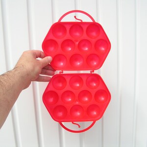 May include: A red plastic egg carrier with a handle. The carrier is open and has 18 compartments for holding eggs.