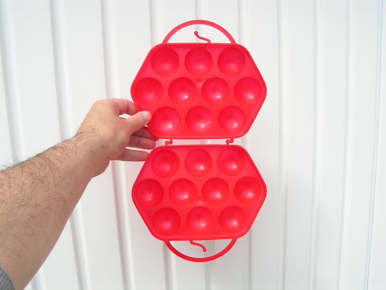 May include: A red plastic egg carrier with a handle. The carrier is open and has 18 compartments for holding eggs.