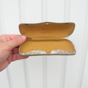 Könnte beinhalten: Ein offenes, rechteckiges Brillenetui in Beige mit gelber Filz-Innenausstattung. Das Etui hat einen silbernen Verschluss.