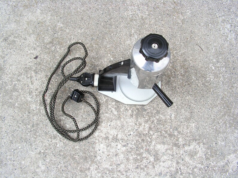 May include: A silver and black electric coffee maker with a black cord and plug. The coffee maker has a round, silver base and a black handle.