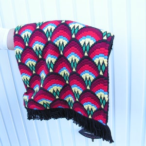 May include: A colorful, patterned textile with a repeating fan-like design in red, burgundy, blue, yellow, and green. The edges are trimmed with black fringe. The textile is held up, showcasing the intricate details.