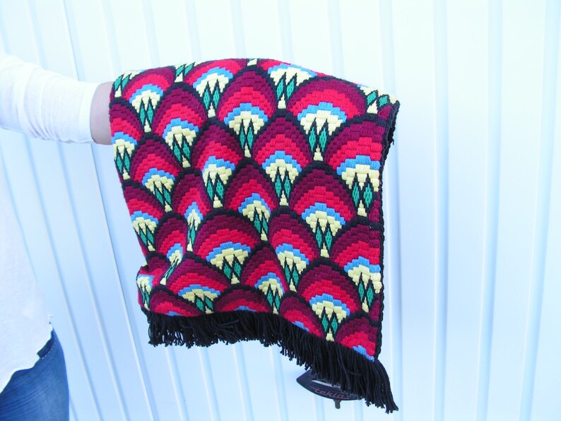 May include: A colorful, patterned textile with a repeating fan-like design in red, burgundy, blue, yellow, and green. The edges are trimmed with black fringe. The textile is held up, showcasing the intricate details.