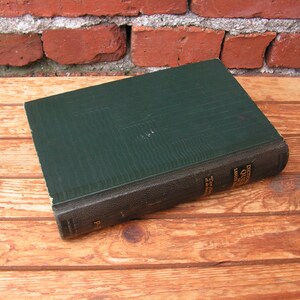 May include: A vintage hardcover book with a dark green cover and a black spine. The book is lying on a wooden surface. The title and author are visible on the spine. The book's cover has a textured appearance.
