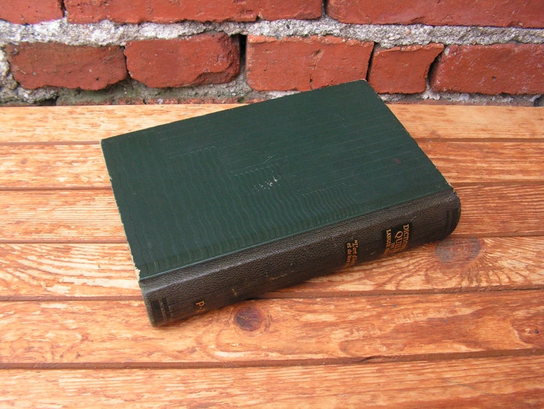 May include: A vintage hardcover book with a dark green cover and a black spine. The book is lying on a wooden surface. The title and author are visible on the spine. The book's cover has a textured appearance.