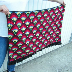 May include: A handmade, rectangular tapestry with a repeating pattern of red, yellow, blue, and black geometric shapes. The tapestry has a black border and fringe along the bottom edge. The tapestry is held up by a person.