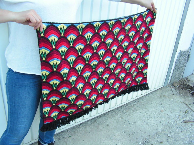 May include: A handmade, rectangular tapestry with a repeating pattern of red, yellow, blue, and black geometric shapes. The tapestry has a black border and fringe along the bottom edge. The tapestry is held up by a person.
