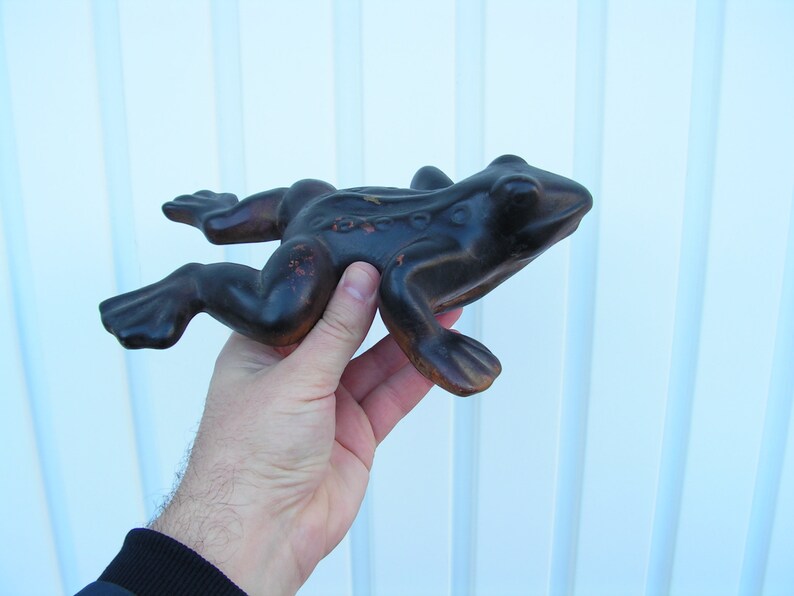 May include: A dark brown cast iron frog figurine. The frog is posed with its legs outstretched and its head tilted slightly to the side. The frog has a textured surface and appears to be hand-painted.