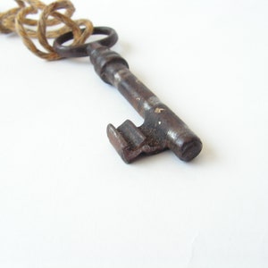 May include: Close-up of a vintage, antique key with a dark, aged metal finish. The key has a detailed design and is attached to a natural-colored rope. The background is plain white, highlighting the key's intricate details.