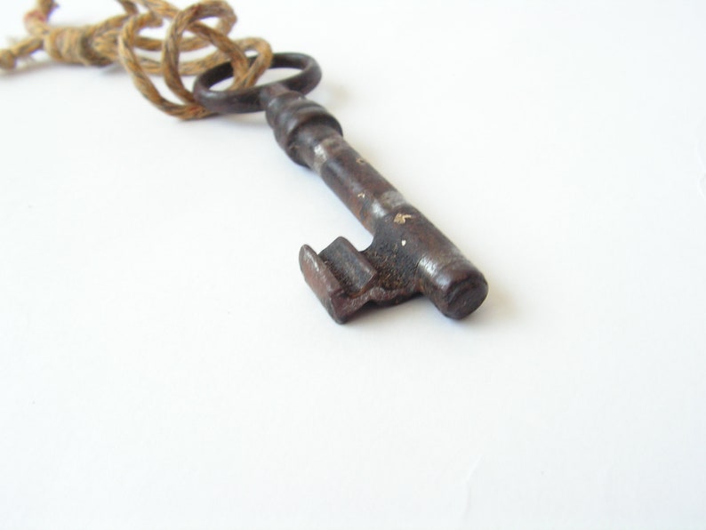 May include: Close-up of a vintage, antique key with a dark, aged metal finish. The key has a detailed design and is attached to a natural-colored rope. The background is plain white, highlighting the key's intricate details.