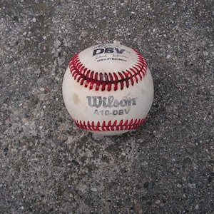 Könnte beinhalten: Ein weißer Wilson A10-DBV Baseball mit roten Nähten. Der Ball zeigt den Text DBV, die Unterschrift des DBV-Präsidenten und den Markennamen Wilson in schwarzer Tinte auf der Oberfläche. Der Baseball weist Gebrauchs- und Abnutzungsspuren auf.