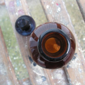 May include: A brown glass bottle with a cork stopper. The bottle is empty and the cork is off. The bottle is sitting on a wooden surface.