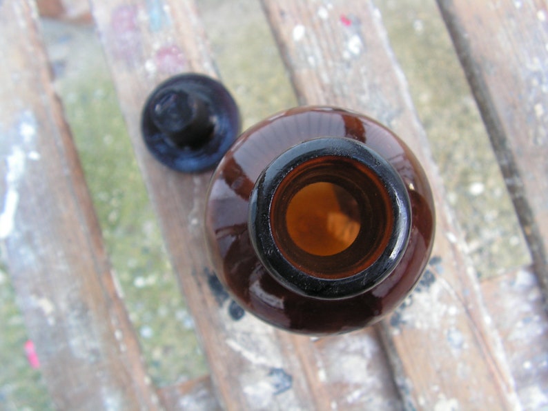 May include: A brown glass bottle with a cork stopper. The bottle is empty and the cork is off. The bottle is sitting on a wooden surface.