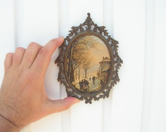 May include: An oval-shaped, antique bronze picture frame with an ornate, openwork border. The framed image depicts a winter scene with figures skating on a frozen pond. The frame is held against a white background.
