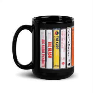 May include: Black ceramic mug featuring cassette tape designs. Tapes include Big Audio Dynamite, The Clash, The Cure, Depeche Mode, and Echo & The Bunnymen. The mug has a large handle and a glossy finish.