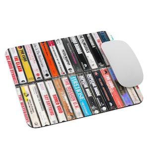 Può includere: Un tappetino per mouse rettangolare con un collage di musicassette vintage con nomi di band come The Cure e Depeche Mode. Un mouse per computer bianco è appoggiato sul lato destro del tappetino, che ha un bordo nero.