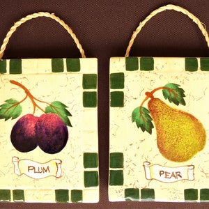 Könnte beinhalten: Zwei Keramikfliesen mit Seilgriffen, jede mit einer anderen Frucht. Die linke Fliese hat einen grün-weißen Mosaikhintergrund mit einer lila Pflaume und dem Wort "PLUM". Die rechte Fliese hat einen grün-weißen Mosaikhintergrund mit einer gelben Birne und dem Wort "PEAR".