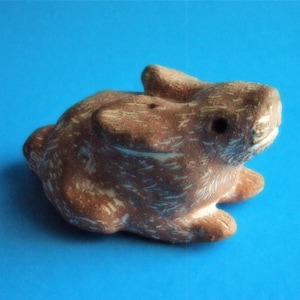 Puede incluir: Una figurita de conejo de cerámica marrón y blanca con una superficie texturizada. El conejo está sentado sobre un fondo azul.