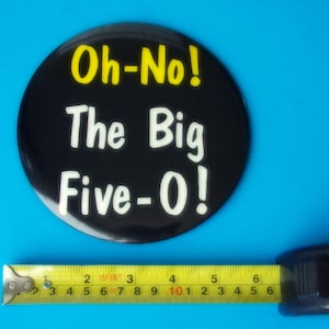 May include: A black round button with white text that reads "Oh-No! The Big Five-O!" The button is on a blue background. A yellow tape measure is in the foreground.