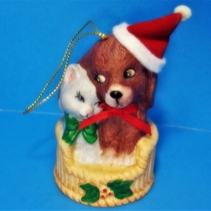 May include: A ceramic Christmas ornament featuring a brown dog wearing a Santa hat and a white cat. The dog and cat are embracing. The ornament is in a basket-like base with a gold string for hanging.