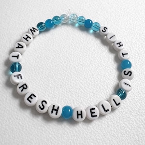 May include: A beaded bracelet with white letter beads spelling "WHAT FRESH HELL IS THIS" and interspersed with blue beads. The bracelet is on a white surface.