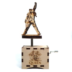 May include: A wooden music box featuring a silhouette of a person with an arm raised in the air. The music box is brown and has a Queen logo on the front. The music box is on a white background.