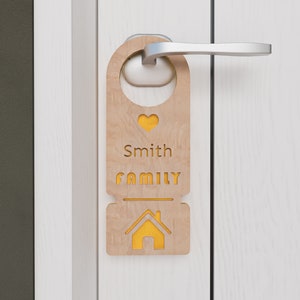 Könnte beinhalten: Ein hölzerner Türhänger mit einem Herzen, dem Text "Smith Family" und einem Haussymbol. Der Hänger ist an einem Türgriff befestigt.