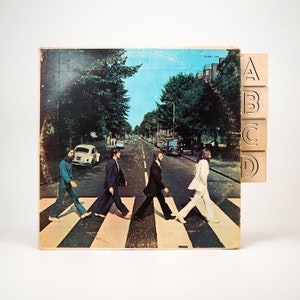 May include: An album cover depicting the Abbey Road crossing, with four wooden blocks labelled A, B, C, and D. The image shows a street scene with a blue sky and trees. The album cover is slightly worn.