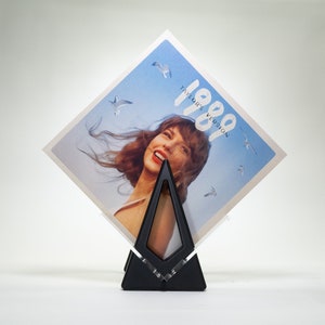 May include: A black vinyl record stand with a clear acrylic insert, holding a vinyl record with a blue sky and white clouds background. The record cover features a woman with long brown hair and the text "1989 Taylor's Version".