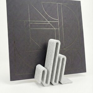 May include: A grey, speckled, abstract sculpture with a folded design stands in front of a dark grey square with gold geometric lines. The sculpture has a unique, modern aesthetic.