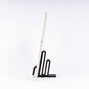 May include: A black metal record stand holding a vinyl record. The stand has a unique, minimalist design with a series of curved lines. The record is off-white with some text visible. The background is plain white.